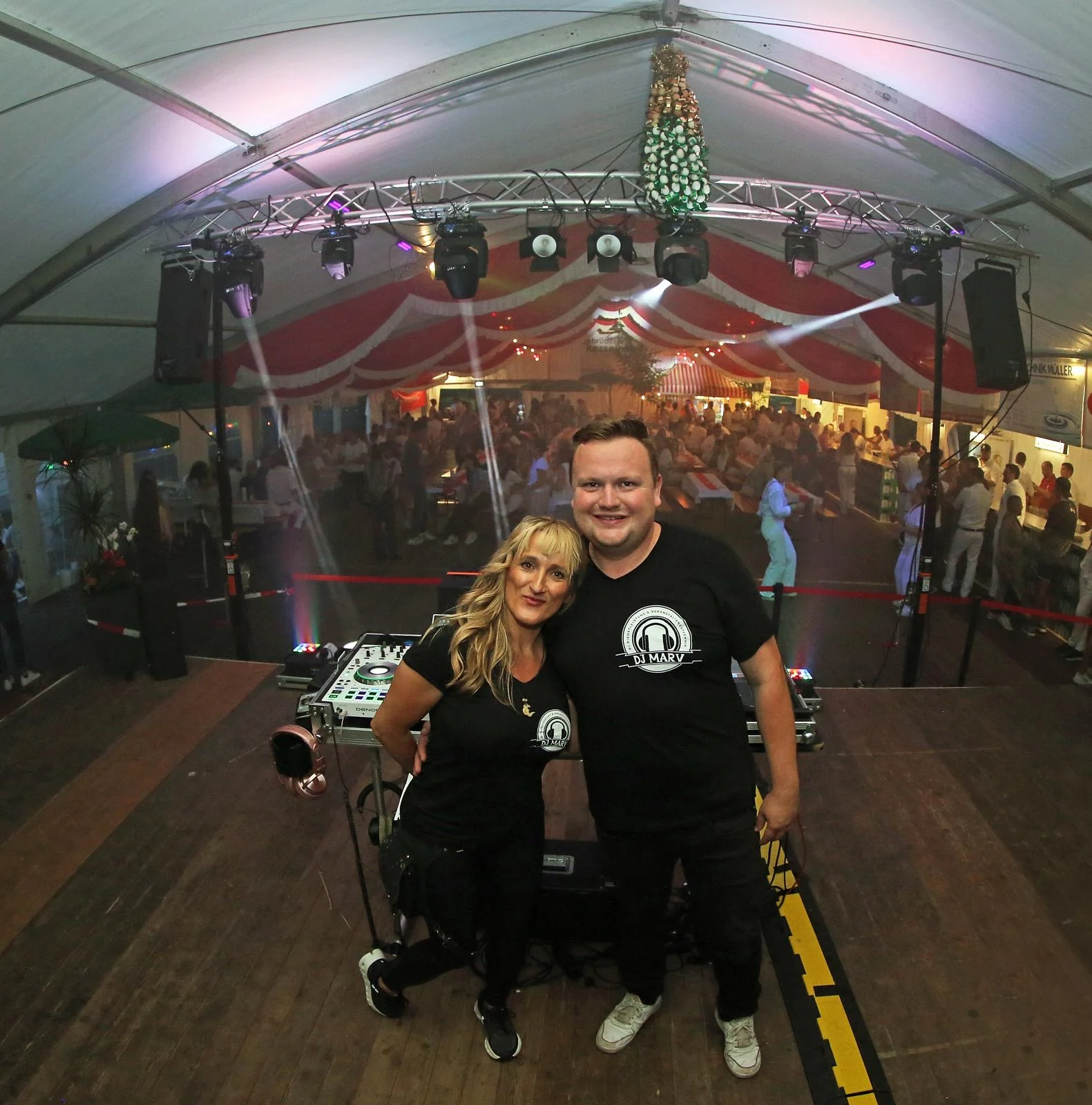 DJ Marv und DJane Sporty Connie auf einer Bühne im Festzelt, vor DJ-Equipment und feierndem Publikum.